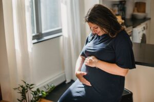 Femeie însărcinată care își ține mâinile pe burtică, lângă o fereastră luminoasă